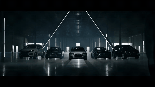 Cinematic concierge walking through a lineup of five Audi vehicles in a dramatic dark showroom with angular lighting