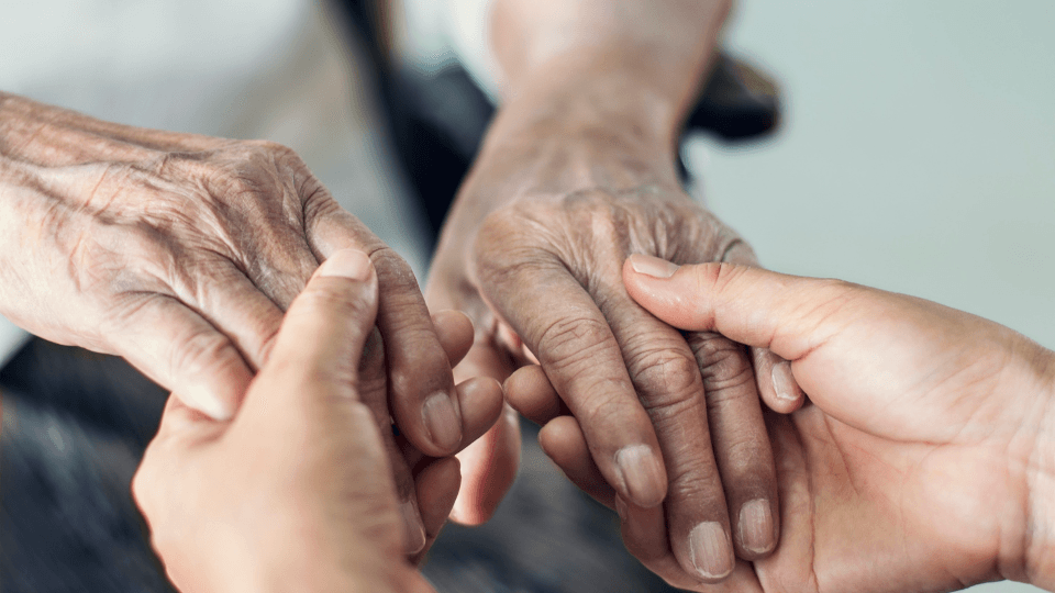 Close-up of a younger person gently holding an elderly person's hands, symbolizing the caregiver-patient bond